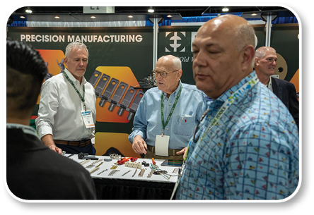 A group of men are standing around a table with a sign that says “precision manufacturing." Description generated by AI